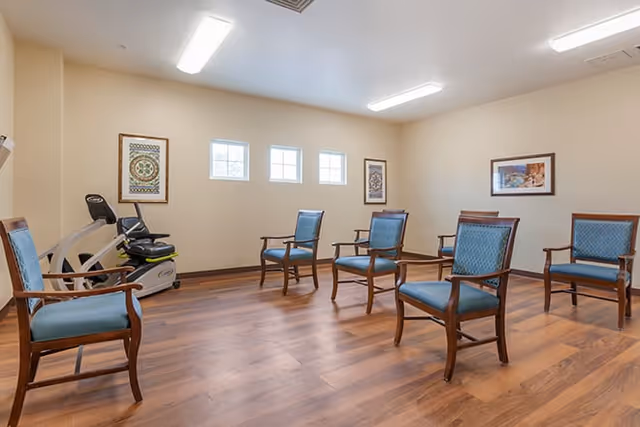 A room with six wooden chairs with blue cushions arranged in a circle on a wooden floor. There is an exercise bike in the corner near a beige wall with three small square windows. The walls have framed artwork, and the ceiling has a rectangular fluorescent light.