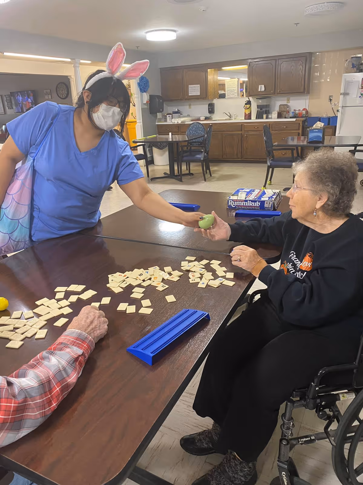 A caregiver wearing bunny ears and a face mask hands a green apple to an elderly woman in a wheelchair. They are sitting at a table with Rummikub game tiles scattered on it in a room with wooden cabinets and chairs.