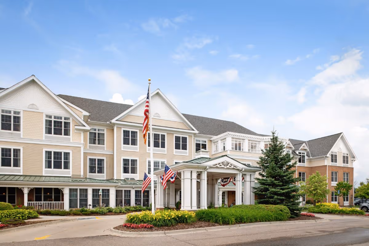 Front exterior of a multi-story senior living building with a covered porte-cochère, landscaped entrance, and American flags.