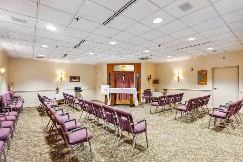 Interior view of a chapel or small worship room with rows of purple cushioned chairs arranged facing an altar. The altar is covered with a white cloth and has a crucifix mounted on the wall behind it. The room has beige walls, carpeted floor with a patterned design, and ceiling with recessed lighting. There are wall sconces providing additional light and a few small tables along the walls.