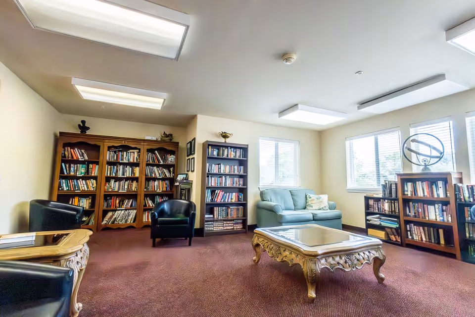 A cozy reading room with multiple bookshelves filled with books, two black armchairs, a light blue couch with a floral pillow, and an ornate wooden coffee table with a glass top. The room has large windows letting in natural light and a maroon carpeted floor.