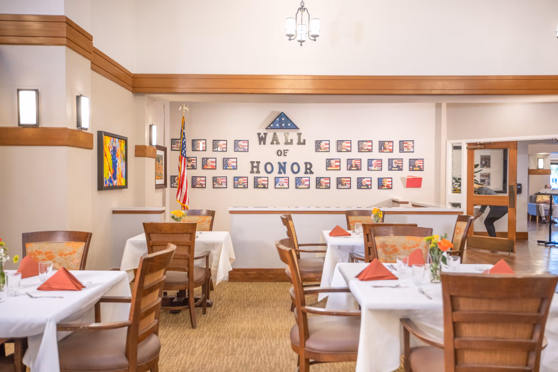 A dining room with several tables covered in white tablecloths, each set with glasses, silverware, and red folded napkins. Wooden chairs with cushioned seats surround the tables. On the back wall is a 'Wall of Honor' display featuring framed photos with American flags and a folded flag in a triangular case above the text. An American flag stands in the corner, and colorful artwork hangs on the left wall. The room is warmly lit with wall sconces and a hanging light fixture.