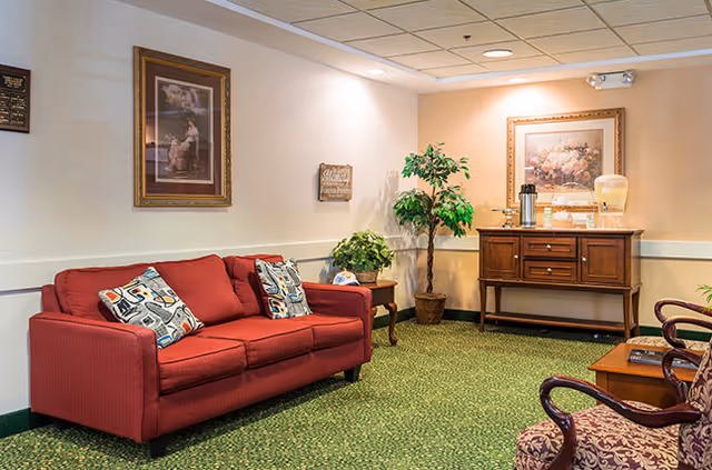 Cozy seating area with a red sofa, patterned chairs, potted plants, framed artwork, and a wooden sideboard with refreshments.