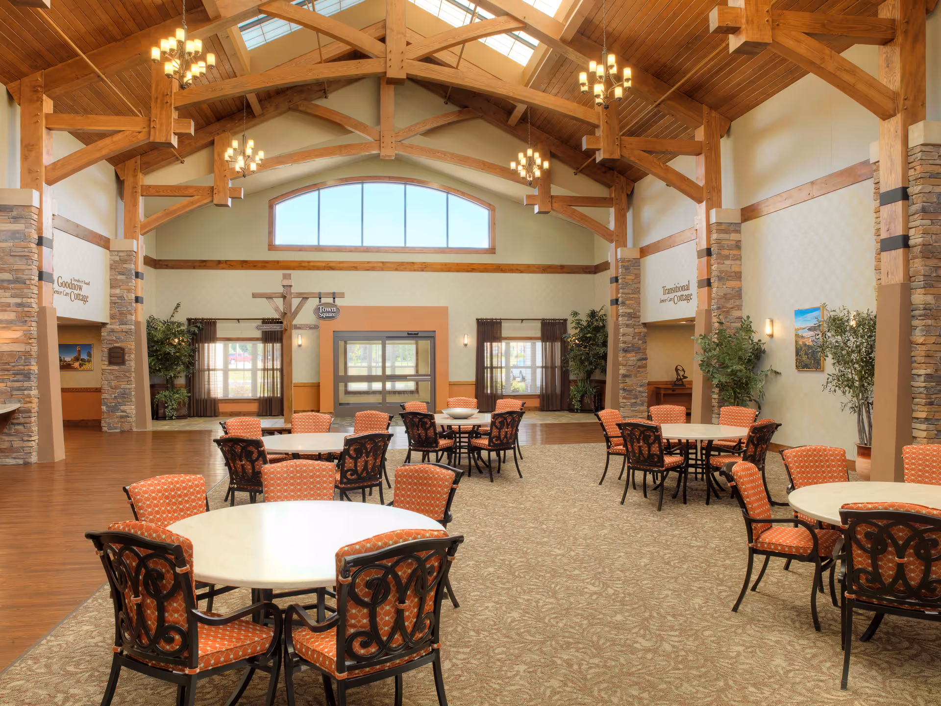 Spacious interior common area with high wooden beam ceiling and large windows. Several round tables with orange patterned chairs are arranged on a carpeted floor. Stone pillars and plants decorate the space, with signs indicating 'Goodnow Senior Care Cottage' and 'Transitional Senior Care Cottage'.
