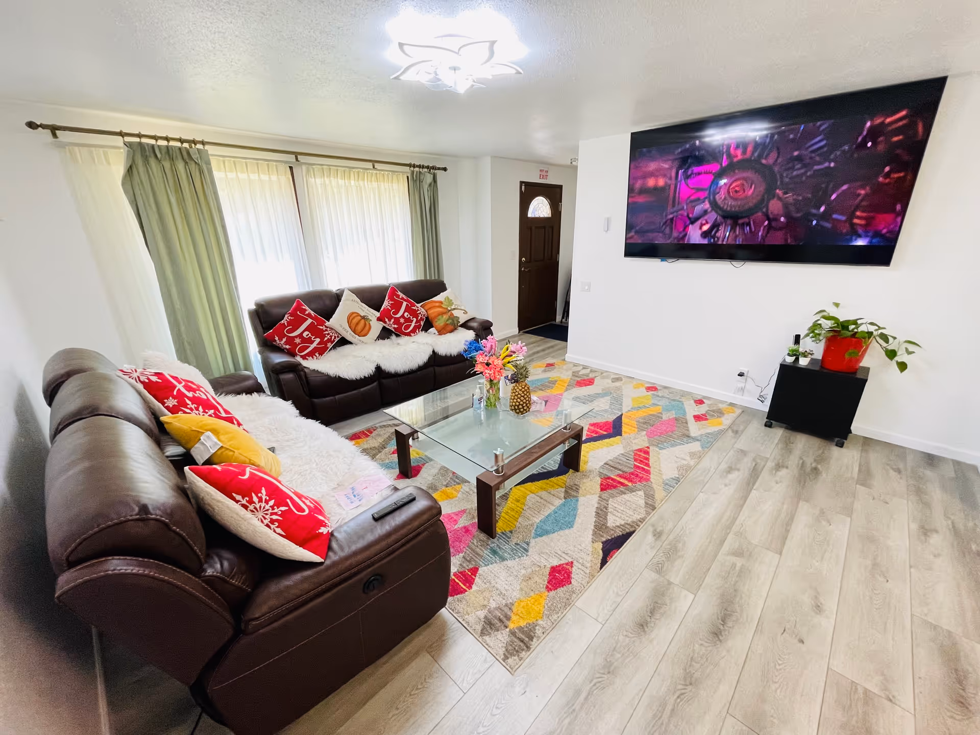 A bright living room with two brown leather sofas adorned with festive red and white pillows and white fluffy throws. A colorful geometric patterned rug covers the floor beneath a glass coffee table with flowers and a pineapple on it. A large flat-screen TV is mounted on the white wall opposite the sofas. There is a small black stand with potted plants next to the TV. Light wood flooring and large windows with sheer curtains and green drapes brighten the room.
