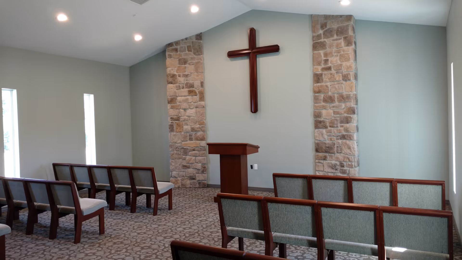 Interior view of a small chapel or worship room with rows of wooden chairs with green cushions facing a wooden podium. The wall behind the podium features two stone columns and a large wooden cross mounted in the center. The room has light gray walls and carpeted floor with a patterned design, and narrow vertical windows on the side walls.