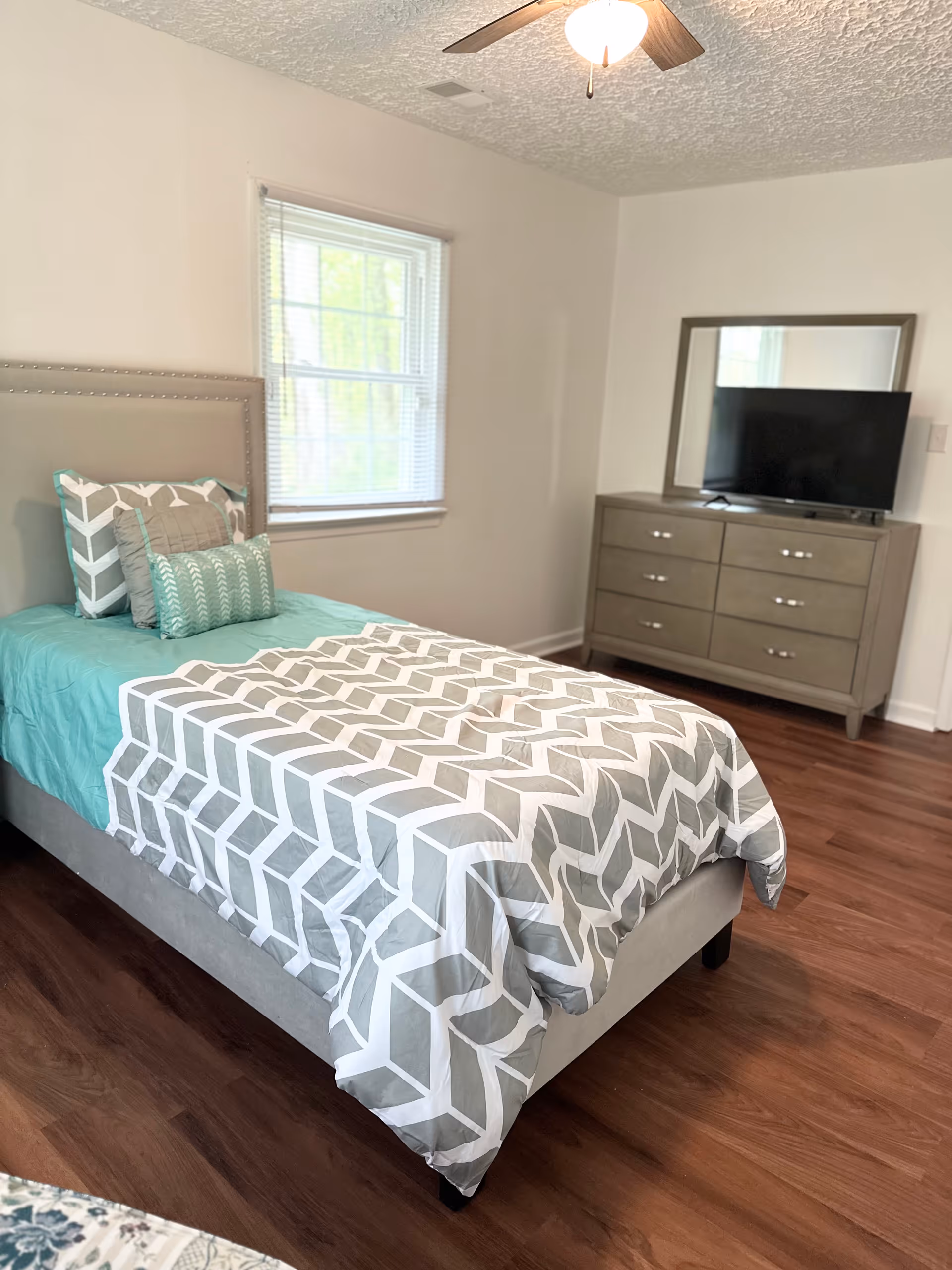 A bedroom with a single bed featuring a gray and white geometric patterned comforter and teal sheets. There are multiple pillows on the bed. A window with blinds is on the wall behind the bed. Across from the bed is a gray dresser with six drawers and a large mirror on top. A flat-screen TV is placed on the dresser. The room has wooden flooring and a ceiling fan with a light.