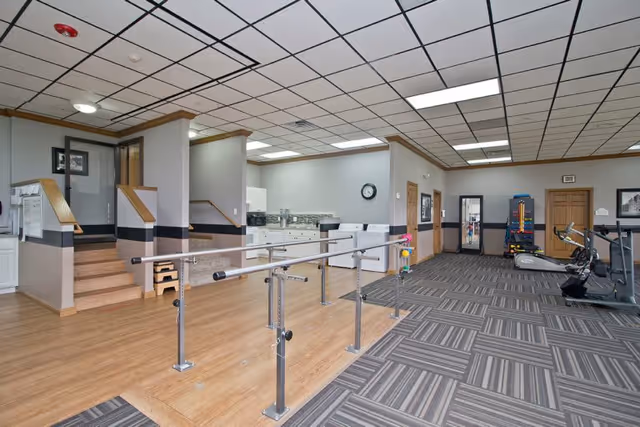Interior view of a senior living facility room with a wooden floor section featuring parallel bars for physical therapy or rehabilitation exercises. The room also has a carpeted area with exercise equipment including a stationary bike and a treadmill. There is a kitchenette area with white cabinets and appliances, and a clock on the wall. The ceiling has a grid pattern with fluorescent lighting.
