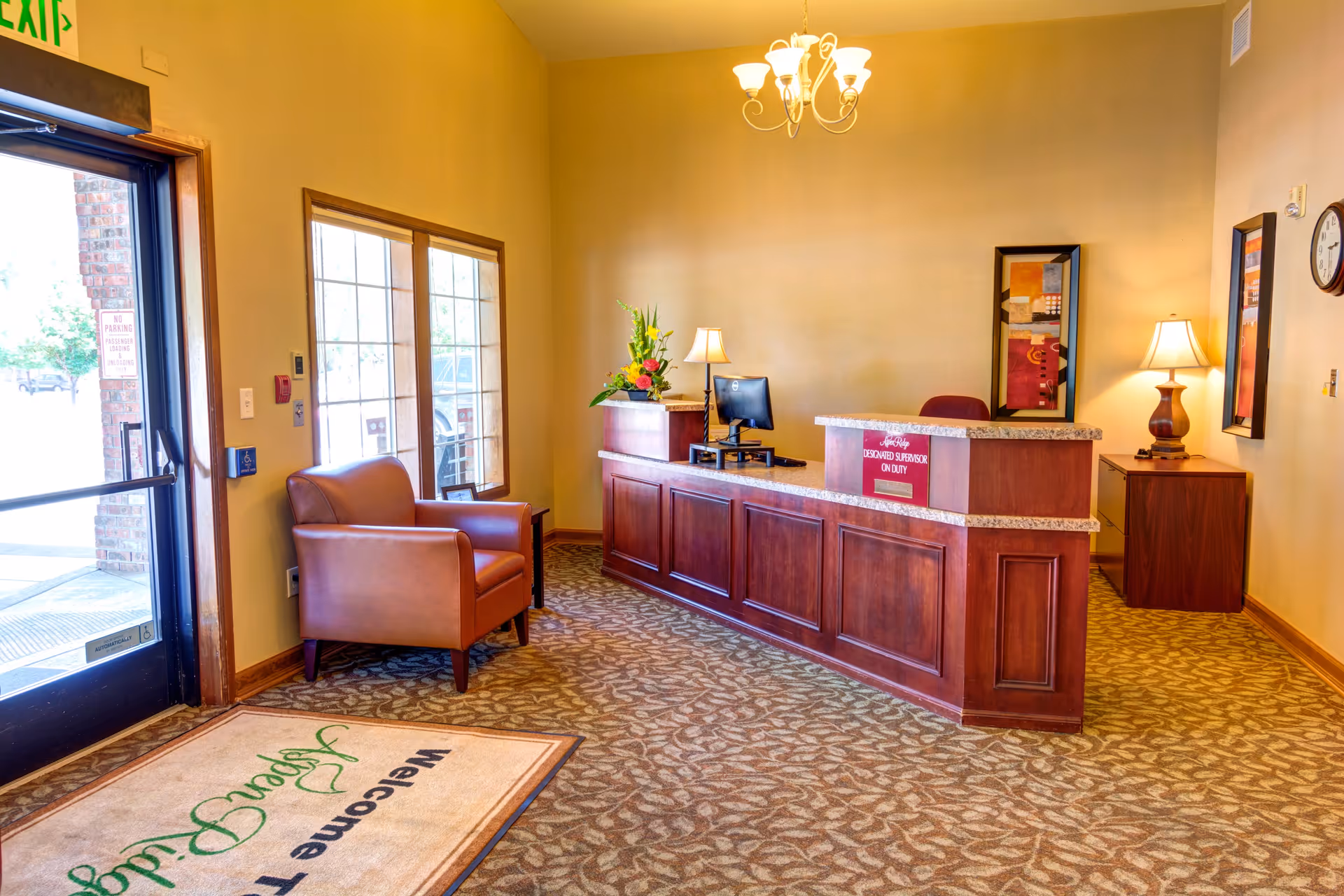 Reception area of Aspen Ridge Memory Care facility with a wooden front desk, a computer, two table lamps, a flower arrangement, a brown leather armchair near a window, and a welcome mat on the carpeted floor. The walls are painted beige and decorated with framed artwork.