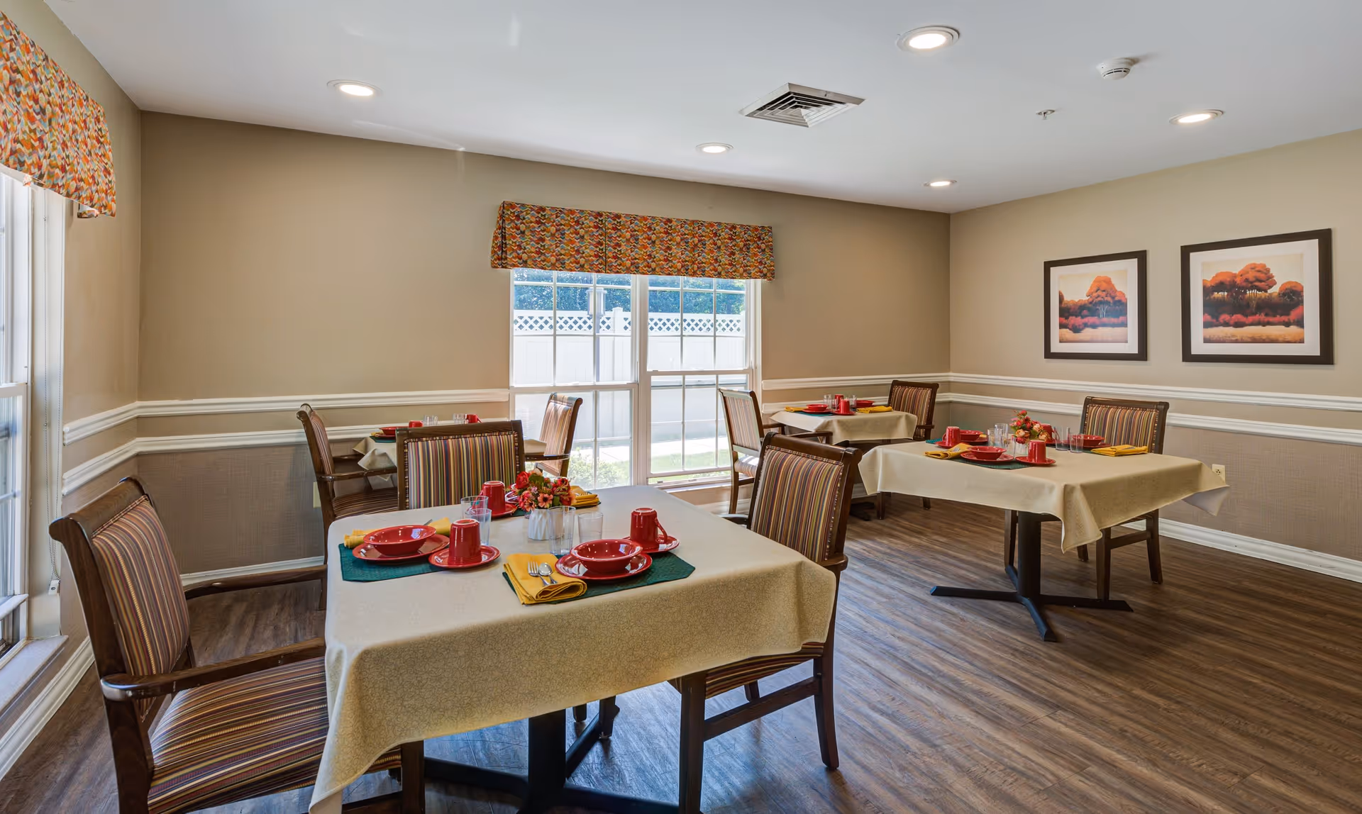 A dining room with four tables, each covered with beige tablecloths and set with red plates, cups, and yellow napkins. The room has wooden flooring, beige walls with white trim, two framed landscape paintings on the wall, and large windows with colorful valances letting in natural light.