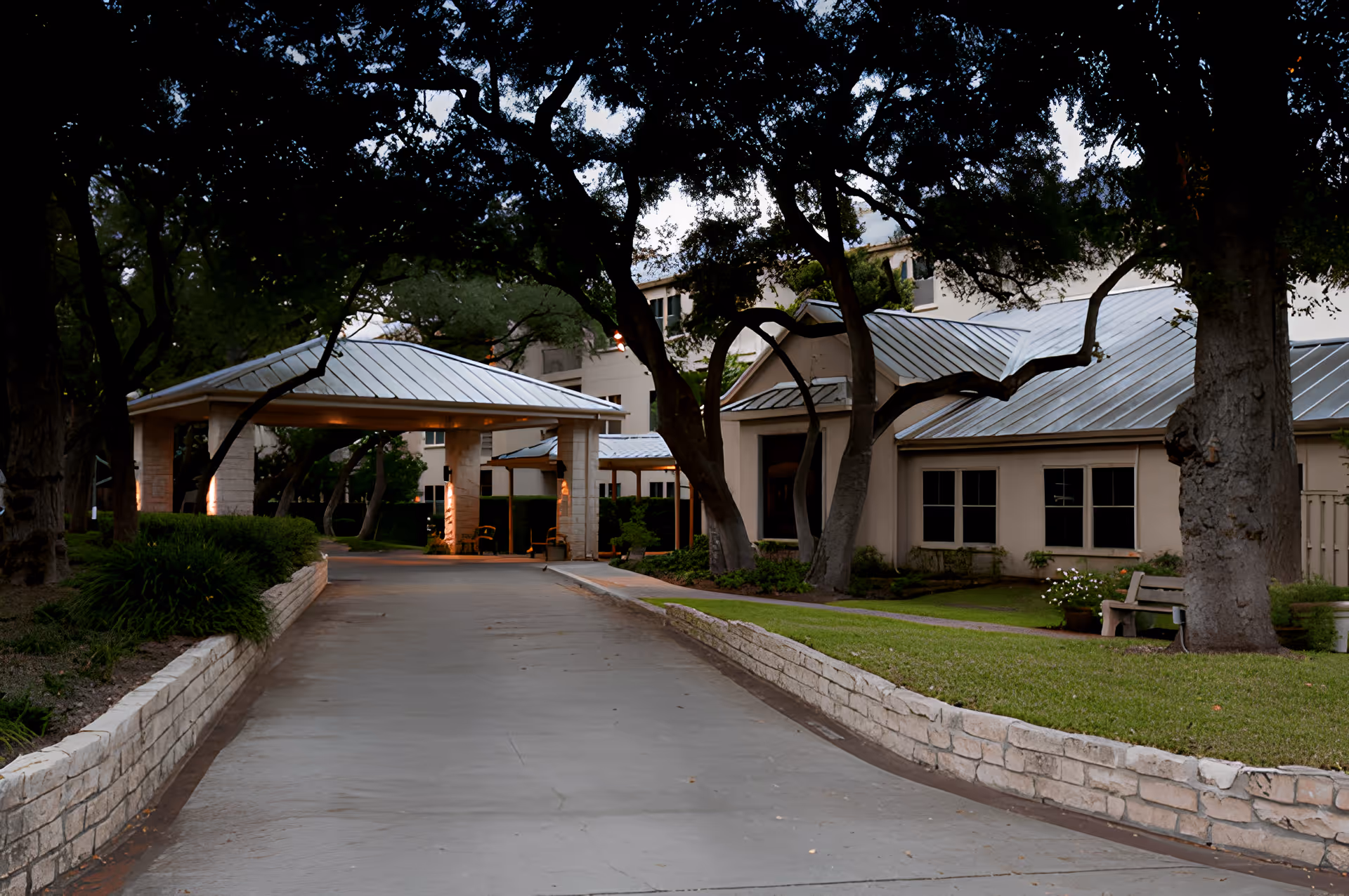 Driveway leading to a covered entrance of a single-story building framed by trees, a low stone wall, and a bench.