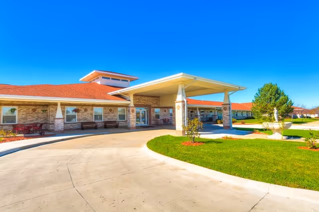 Exterior view of a single-story senior living facility with a covered entrance, brick walls, red roof, and a circular driveway. There is a green lawn with small trees and a decorative fountain statue near the entrance under a clear blue sky.