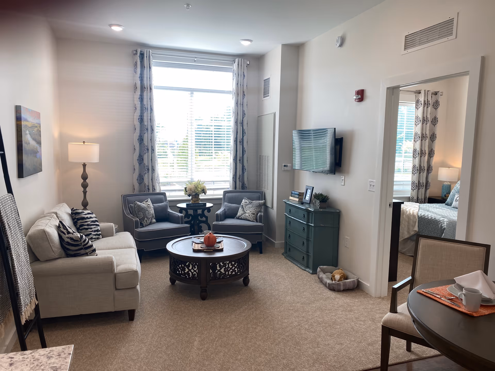 Well-lit living room with a sofa, two armchairs around a round coffee table, a wall-mounted TV and a view into a bedroom.