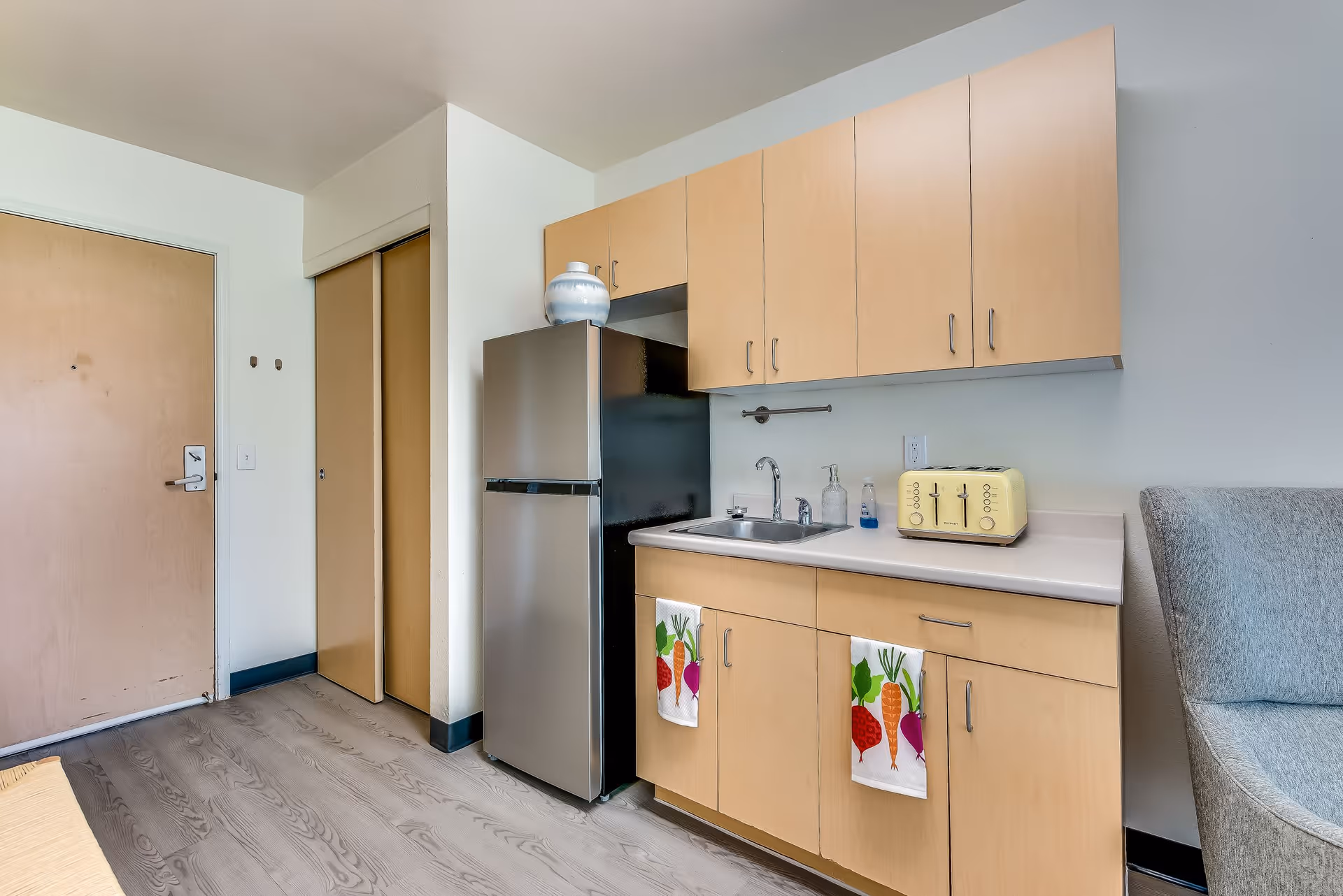 A small kitchenette area in a senior living facility with light wood cabinets, a stainless steel refrigerator, a sink, a yellow toaster, and two colorful vegetable-themed hand towels hanging from the cabinet doors. To the left is a closed wooden door and a closet with sliding doors. Part of a gray upholstered chair is visible on the right.
