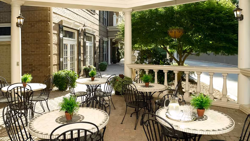 Outdoor patio area with round mosaic tile tables and black metal chairs. Each table has a small potted green plant. There is a pitcher of water and glasses on one table. The patio is adjacent to a brick building with large windows and white columns, and there is greenery including a tree and bushes surrounding the area.