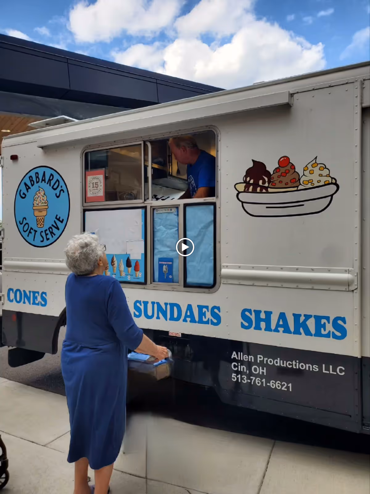 An elderly woman in a blue dress stands outside an ice cream truck labeled 'Gabbard's Soft Serve,' ordering from a man inside the truck. The truck has illustrations of ice cream cones and sundaes, and text advertising cones, sundaes, and shakes. The sky is partly cloudy in the background.