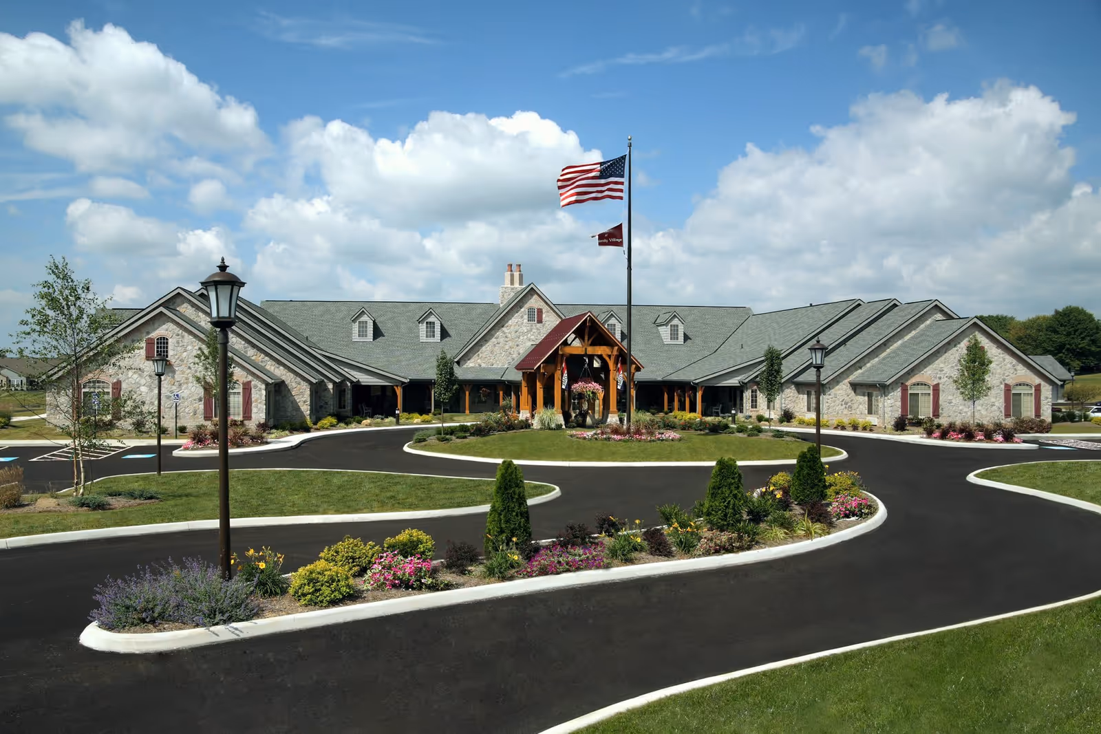 Front exterior of a single-story stone building with a circular driveway, landscaped islands, and flagpoles under a blue sky.