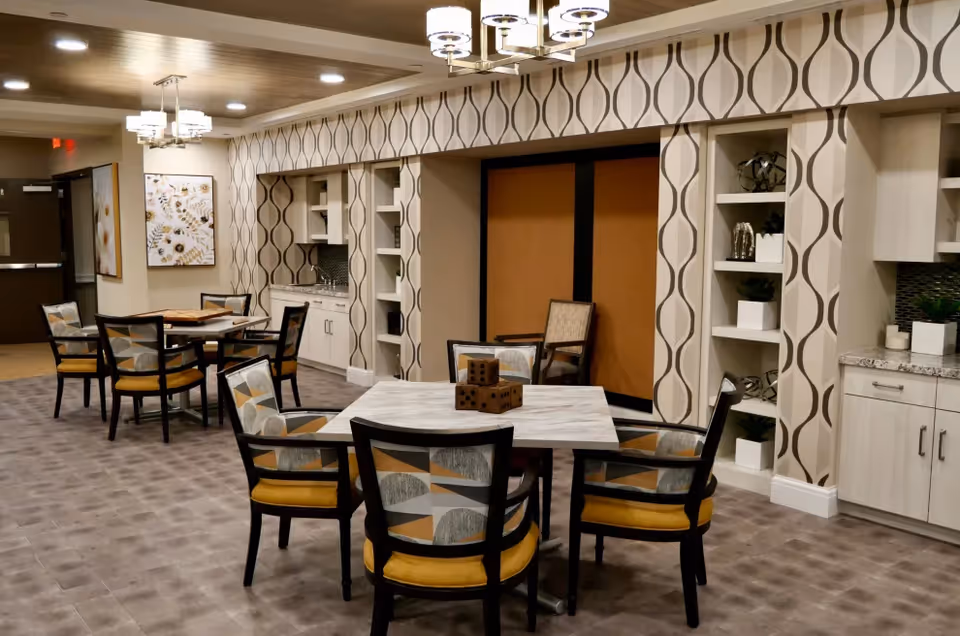 A well-lit interior room with multiple square tables surrounded by chairs with patterned upholstery and yellow cushions. The walls feature a modern geometric wallpaper design, built-in shelves with decorative items, and a small kitchenette area with cabinets and a sink. Ceiling lights hang above the tables, and framed artwork is visible on the far wall.