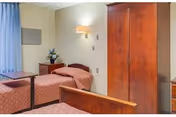 Shared bedroom with two twin beds, a wooden wardrobe, bedside table with lamp, overbed table, and a curtained window.