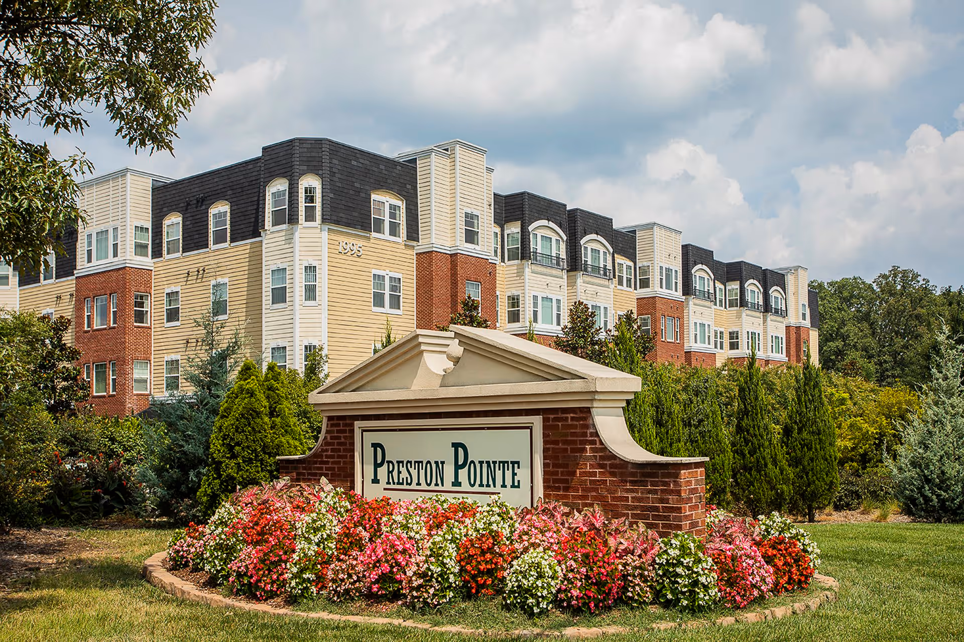 Exterior view of a multi-story residential building with a combination of brick and light-colored siding. In front of the building is a landscaped area with colorful flowers and a brick sign that reads 'Preston Pointe'. Trees and shrubs surround the area under a partly cloudy sky.
