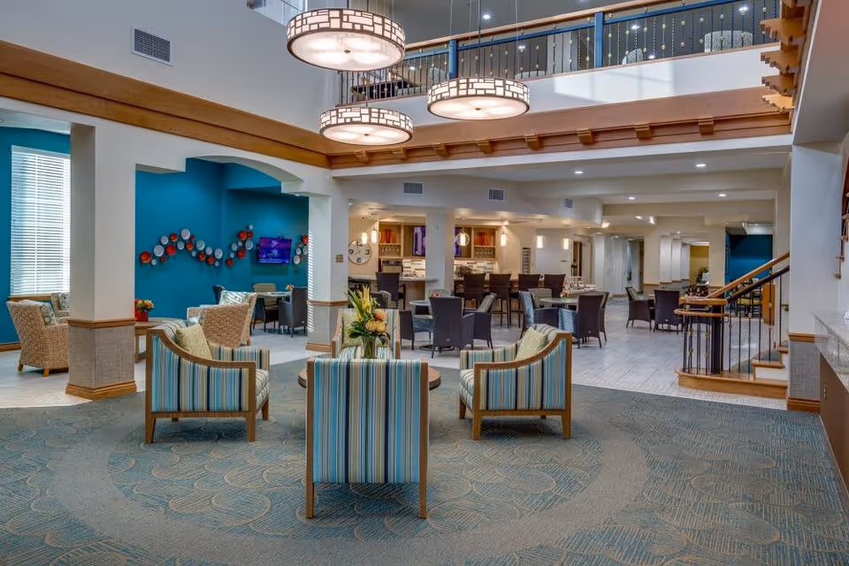 Spacious and well-lit senior living facility common area with comfortable seating including striped armchairs and wicker chairs, a teal accent wall with decorative plates, a wall-mounted TV, and a dining area with tables and chairs. The space features large hanging light fixtures, a staircase with wooden handrails, and a carpeted floor with a patterned design.