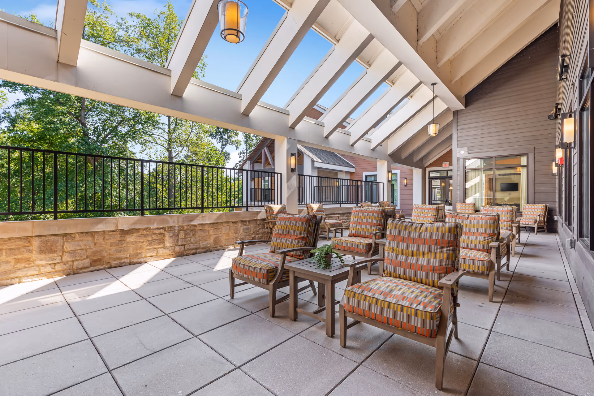 Covered outdoor patio area with multiple cushioned armchairs arranged in rows facing forward, small tables with plants between some chairs, stone half-wall with black metal railing, and a slanted roof with skylights allowing natural light. Trees and blue sky are visible outside the railing.