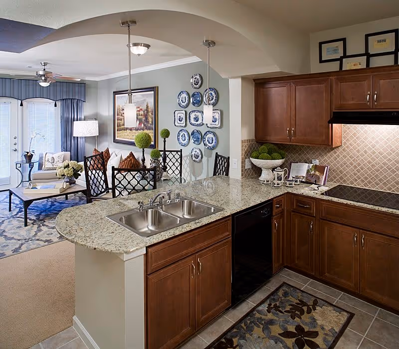 View of a modern kitchen with granite countertops, a double sink, wooden cabinets, and a tiled backsplash. The kitchen opens into a living area with a sofa, armchair, coffee table, and decorative wall plates. Pendant lights hang over the kitchen counter, and a ceiling fan is visible in the living room.