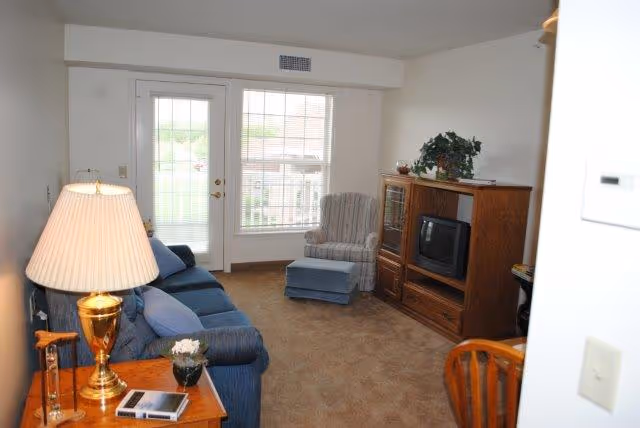Cozy living room with a blue sofa, upholstered armchair and ottoman, wooden TV cabinet, and a glass door with blinds letting in natural light.