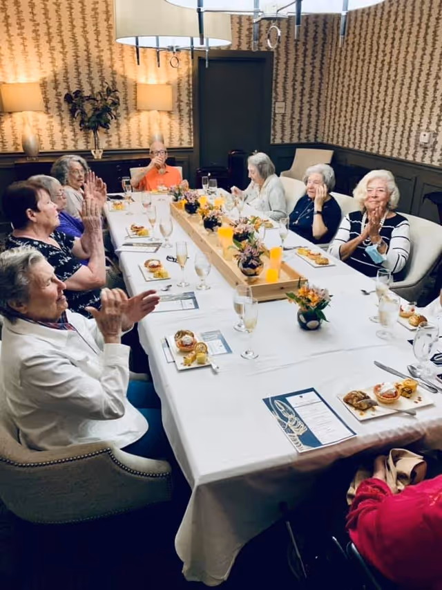 A group of elderly women sitting around a long dining table in a warmly lit room, clapping and enjoying a meal together. The table is set with plates of food, glasses, menus, and decorative flower arrangements in the center. The room has patterned wallpaper, two wall lamps, and a large overhead light fixture.