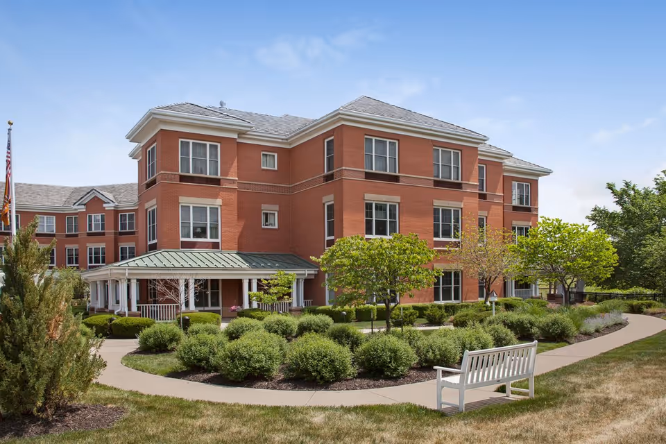 Exterior view of a three-story brick building with multiple windows, surrounded by well-maintained landscaping including bushes, small trees, and a curved walkway. There is a white bench on the grass near the walkway and a flagpole with an American flag on the left side of the image under a clear blue sky.