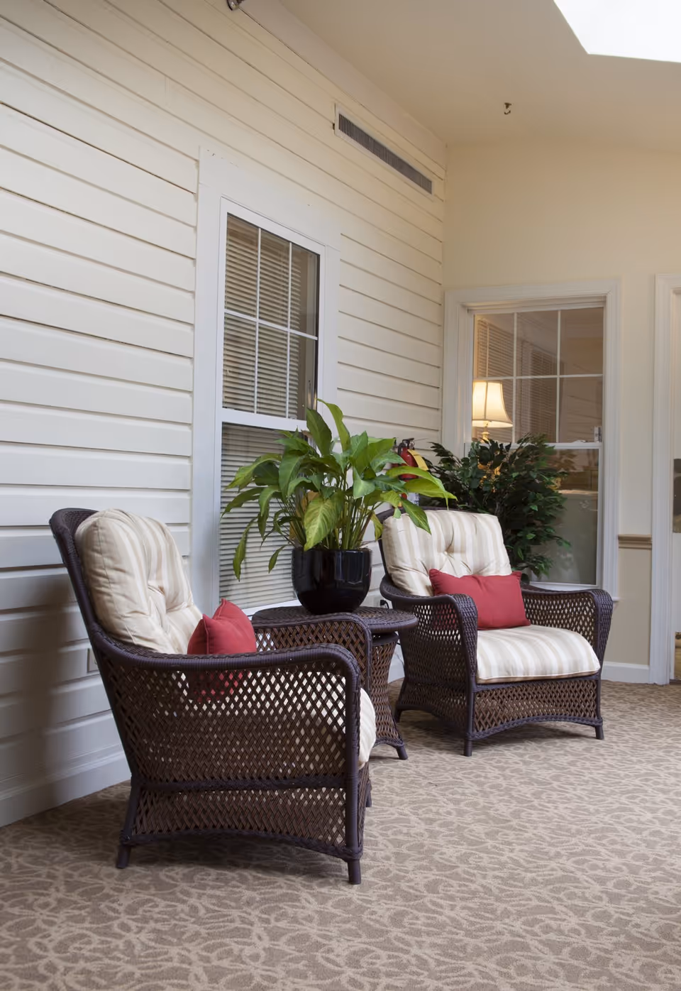 Two cushioned wicker chairs with striped beige cushions and red accent pillows are placed on a patterned carpeted floor in an indoor seating area. A small wicker table with a potted green plant sits between the chairs. The background features cream-colored walls with white trim, a window with blinds, and a door with a glass panel showing a lit lamp and another plant inside.