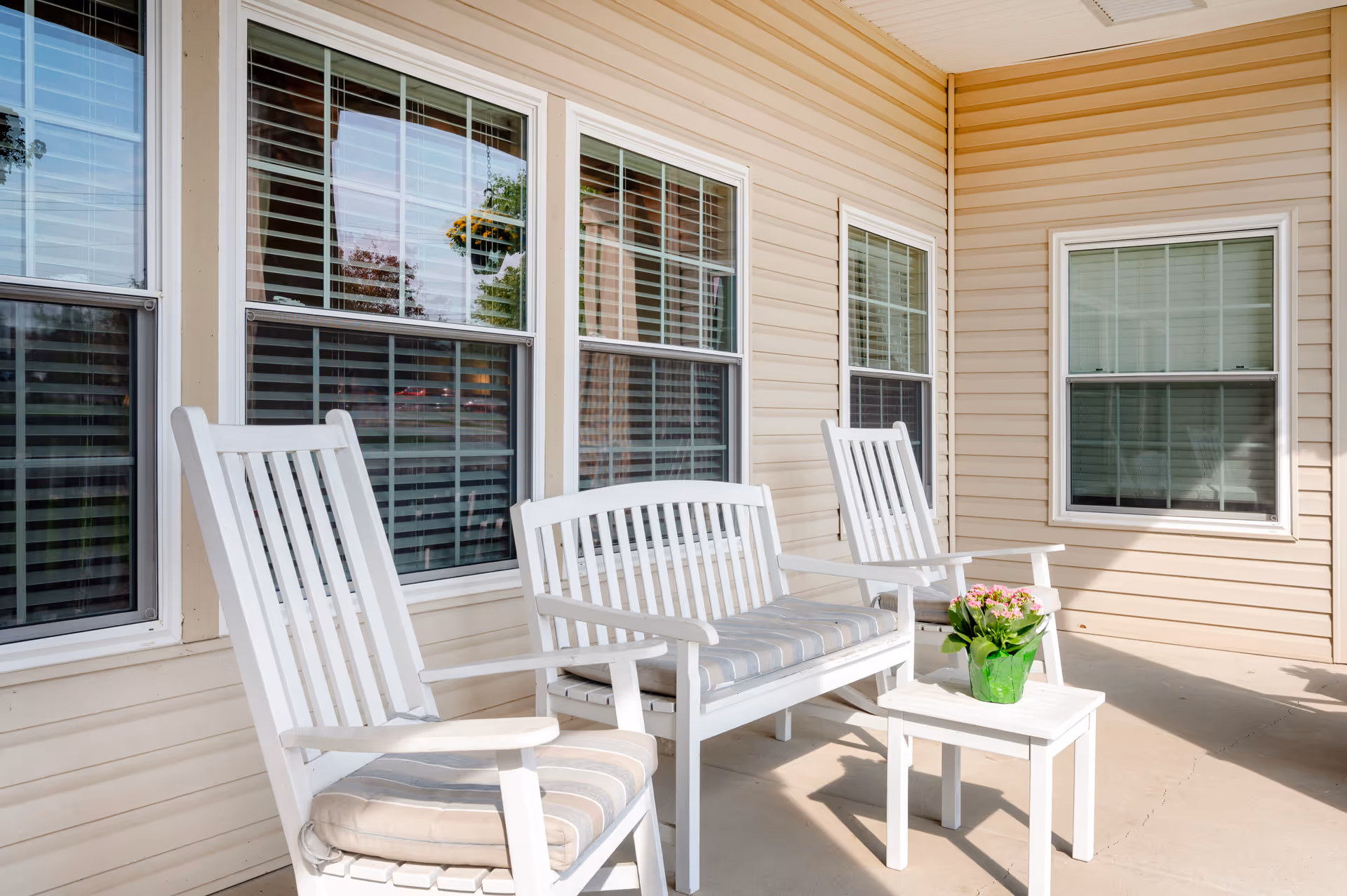 A covered outdoor patio area with beige siding walls and four windows with white frames and blinds. The patio is furnished with white wooden rocking chairs and a bench with striped cushions, and a small white table holding a green pot with pink flowers.