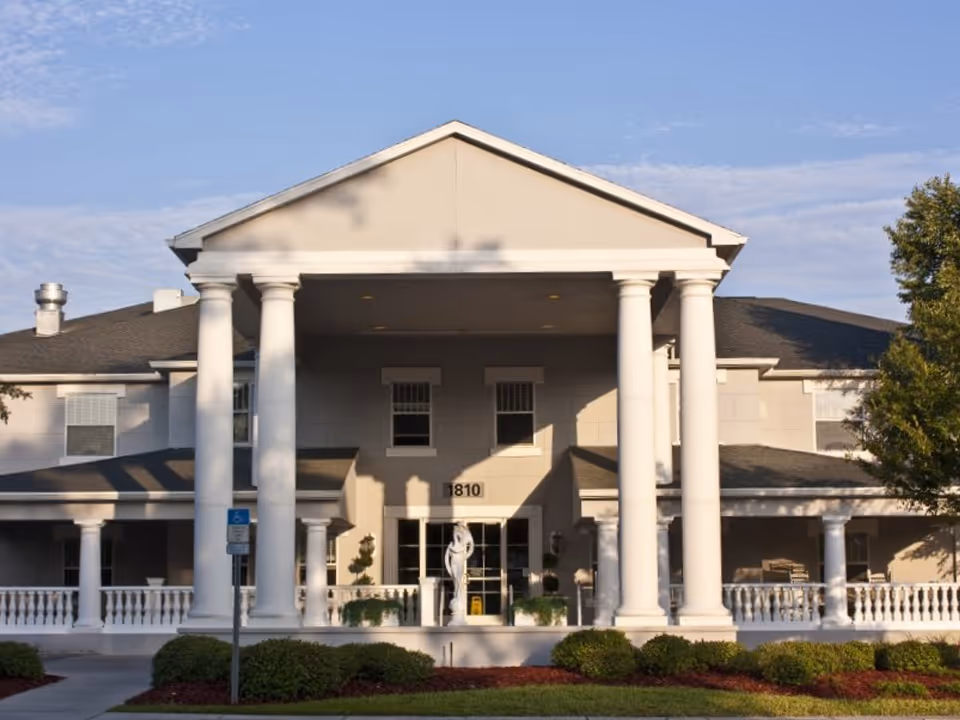Front exterior of a two-story assisted living building with large white columns, central entrance, statue, and landscaped shrubs.