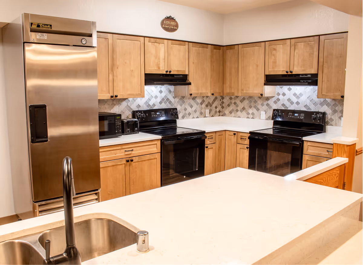 Bright assisted-living kitchen with wooden cabinets, a stainless refrigerator, two black stoves/ovens, tiled backsplash and a light-colored island with a sink.