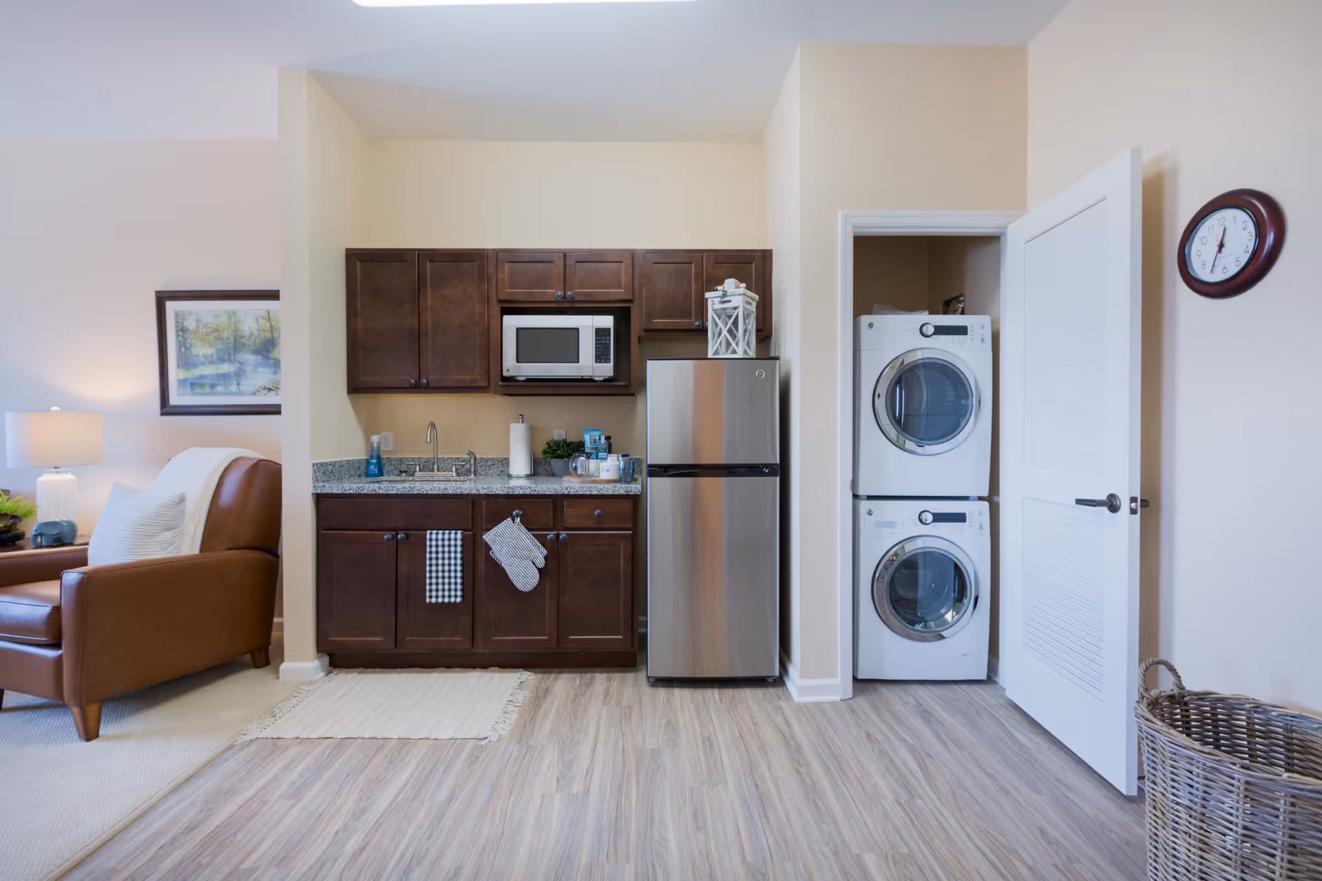A small kitchen area with dark wooden cabinets, a granite countertop, a microwave, and a stainless steel refrigerator. To the right, there is a closet with a stacked washer and dryer behind a partially open white door. A round wall clock is mounted above the door, and a wicker laundry basket is on the floor nearby. To the left, part of a living room is visible with a brown leather armchair, a lamp, and a framed painting on the wall.