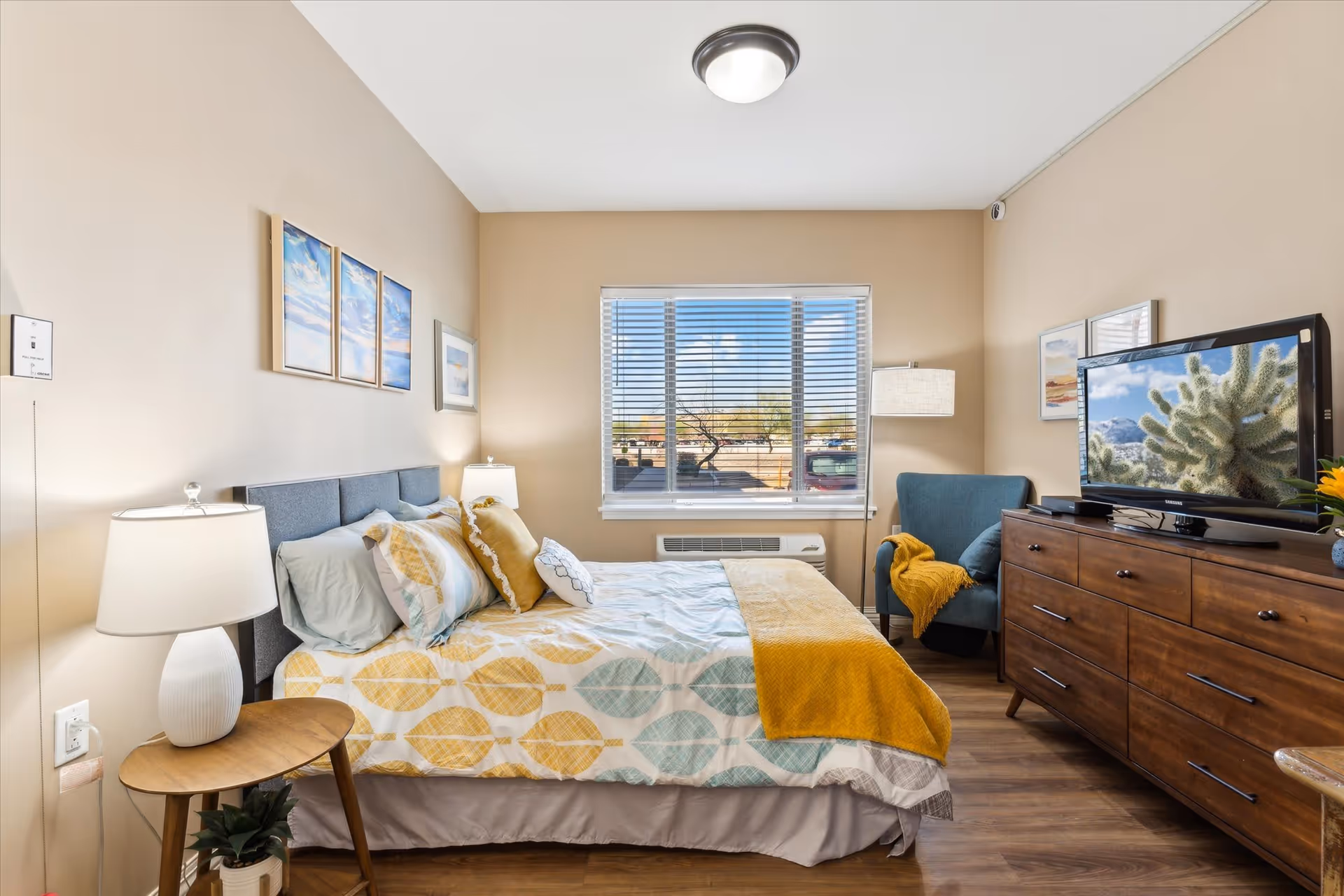 A cozy bedroom with a bed featuring a patterned bedspread in yellow and blue tones, multiple pillows, and a yellow throw blanket. There is a wooden nightstand with a white lamp and a small plant. A large window with blinds lets in natural light, and a blue armchair with a yellow throw is positioned near the window. A wooden dresser with a flat-screen TV displaying a cactus image is on the right side of the room. The walls are decorated with framed artwork.