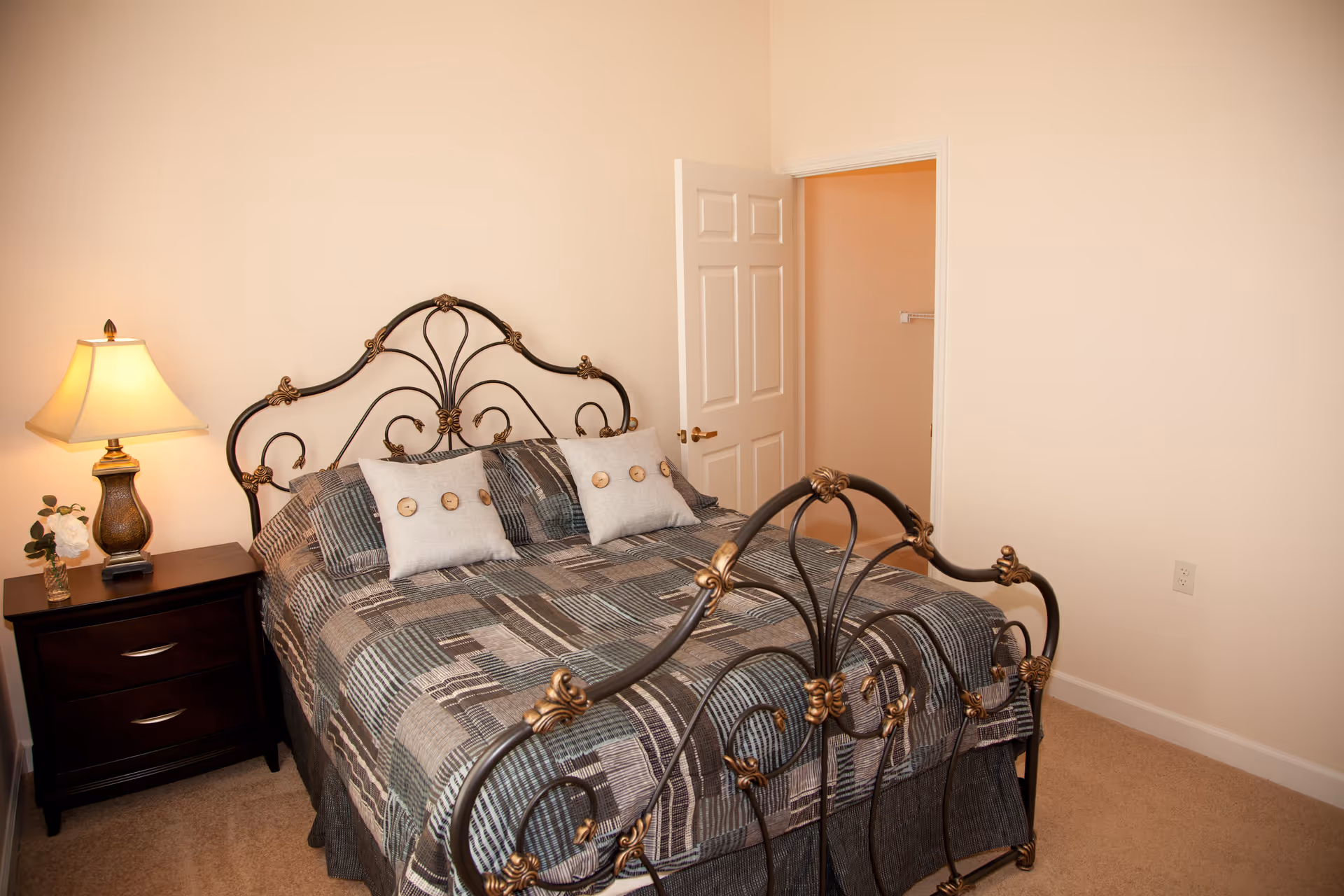 A bedroom with a decorative metal bed frame featuring gold accents, a bed covered with a patterned quilt and three pillows, a dark wooden nightstand with two drawers, a table lamp with a beige shade, and a small flower arrangement. The room has beige walls and carpet, and an open door leading to a closet.