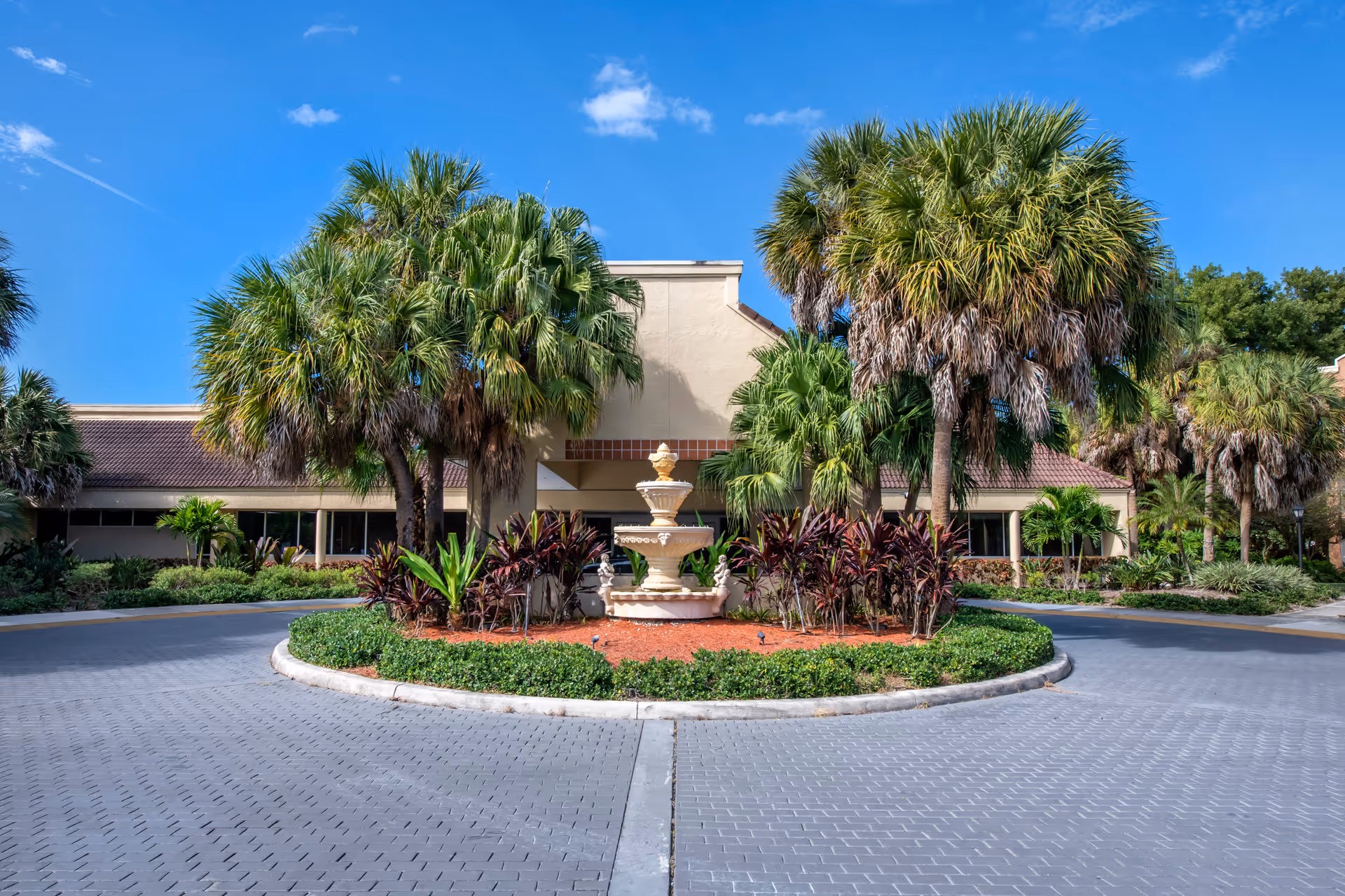 Front exterior view of Brookdale West Palm Beach facility with a circular driveway, landscaped greenery, palm trees, and a decorative fountain in the center under a clear blue sky.