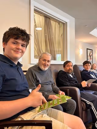 A group of people sitting in a row of chairs inside a well-lit room. The group includes a young boy giving a thumbs up, an elderly man with gray hair and beard, a middle-aged man with a beard, and two other individuals further down the row. The room has light-colored walls, a window with curtains, and framed artwork on the wall.