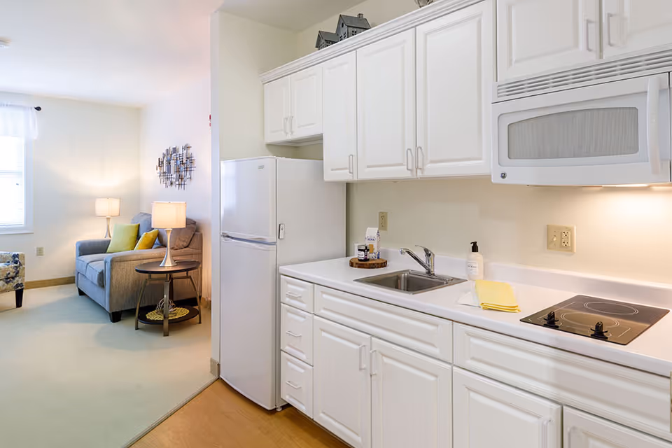 Bright kitchenette with white cabinets, sink, refrigerator and stovetop, opening to a small living area with an armchair and lamps.