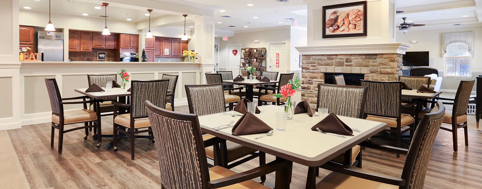 Bright communal dining room with multiple set tables and chairs and an open kitchen/counter in the background.