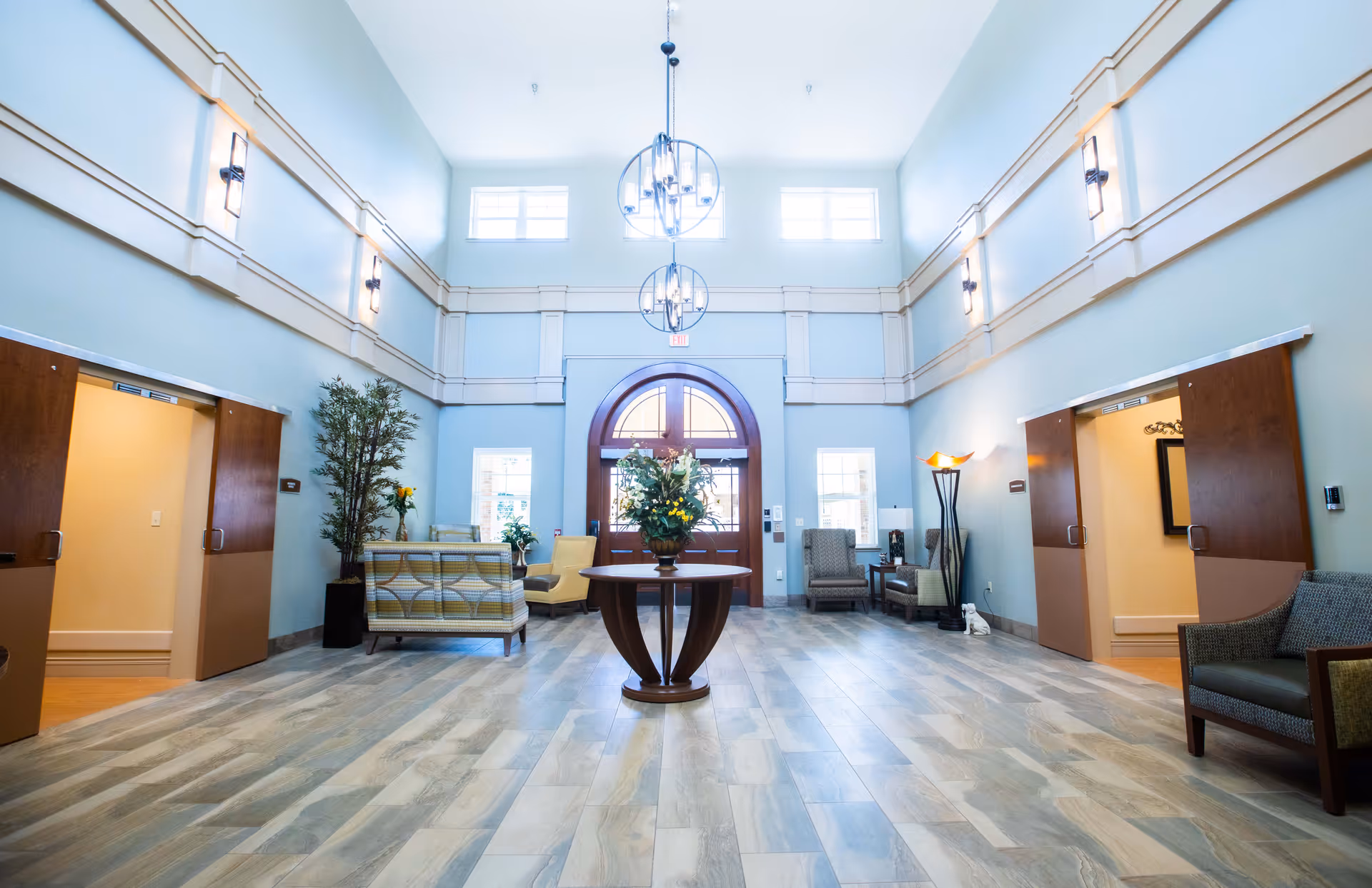 Spacious and well-lit common area with high ceilings, light blue walls, and large windows. The room features a round wooden table with a floral arrangement in the center, several armchairs and sofas arranged along the walls, modern hanging light fixtures, and two open doorways with wooden sliding doors on either side.