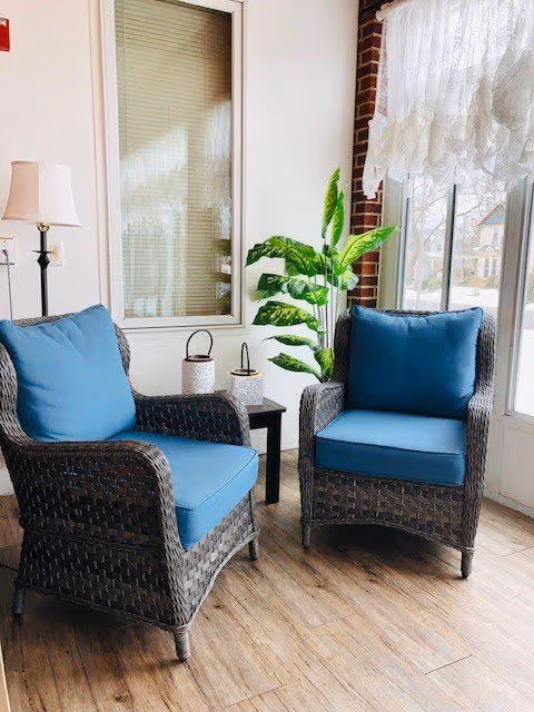 Two wicker armchairs with blue cushions placed on a wooden floor next to a window with white lace curtains. A small black side table with two decorative lanterns sits between the chairs. A tall green plant is positioned near the window, and a floor lamp with a white shade is visible in the corner.