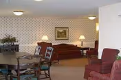 Community lounge with maroon sofas and armchairs, side tables and lamps, patterned wallpaper, and a small dining table in the foreground.