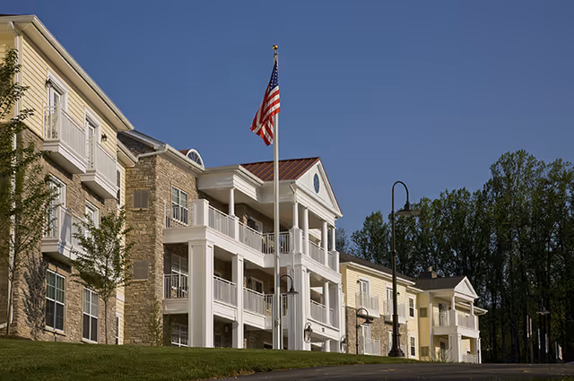 Exterior view of a senior living facility building with multiple balconies, white columns, and an American flag on a flagpole in front. The building has a combination of stone and yellow siding, with trees in the background under a clear blue sky.