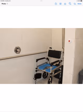 A shower area with beige tiled walls featuring a metal grab bar and a shower control knob. There is a blue and black shower wheelchair positioned inside the shower space, designed for accessibility.
