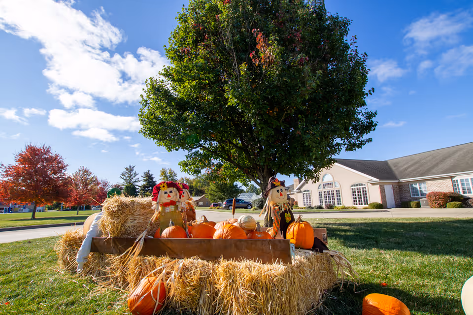 Outdoor autumn scene at a senior living facility with hay bales, pumpkins, and two scarecrow decorations under a tree, with a building and colorful fall trees in the background under a partly cloudy blue sky.