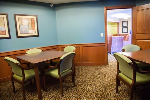 Small dining/activity room with two wooden tables, green-upholstered chairs, teal walls and patterned carpet, opening into a lounge through a doorway.