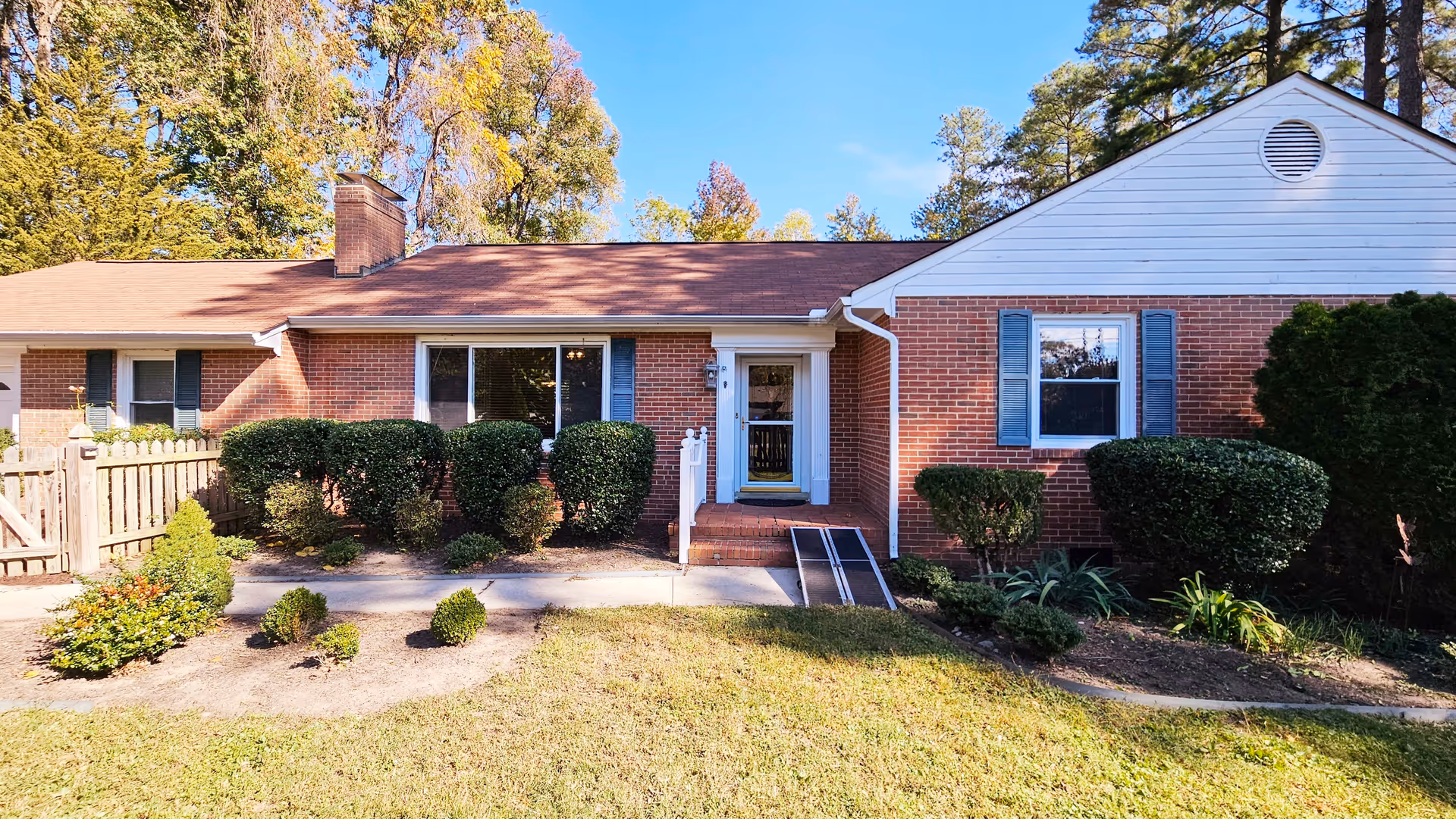 Front exterior view of a single-story brick building with a brown roof, white trim, and blue shutters. There is a small porch with steps and a wheelchair ramp leading to a white door. The front yard has neatly trimmed bushes, a wooden fence on the left, and trees in the background under a clear blue sky.