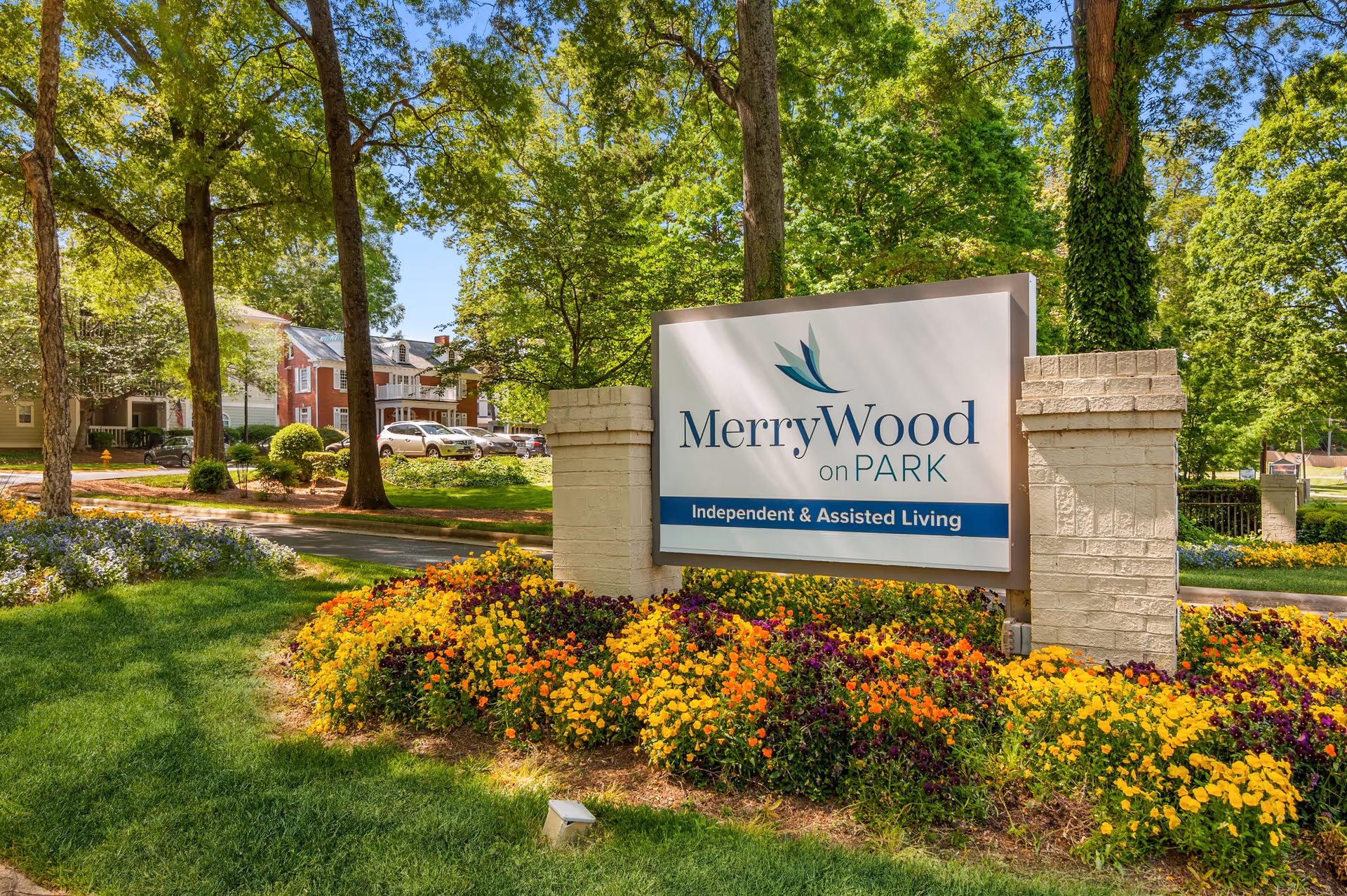 Outdoor view of the MerryWood on Park sign surrounded by colorful flowers and green trees, with residential buildings and parked cars in the background under a clear blue sky.